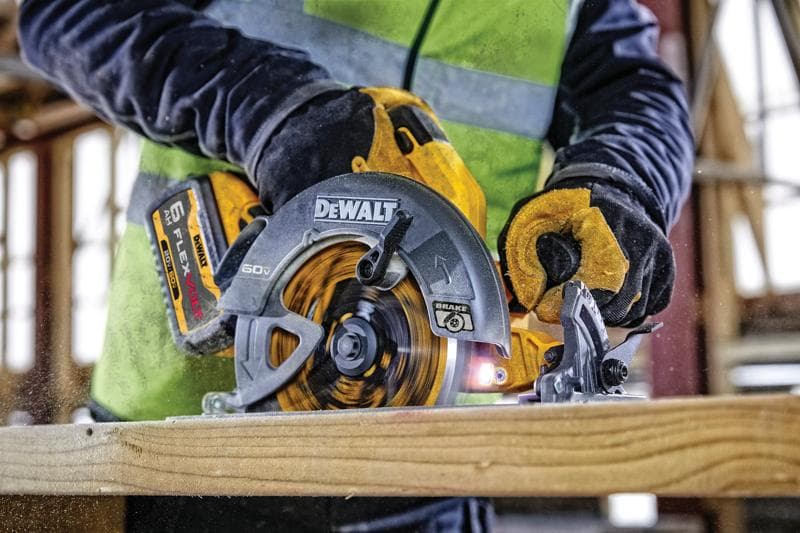 Vista de perto de uma pessoa usando uma serra circular sem fio DEWALT 60V FLEXVOLT para cortar um pedaço de madeira. A pessoa está usando luvas de trabalho e um colete de alta visibilidade em um canteiro de obras.
