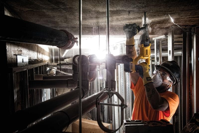 Uma pessoa usando equipamentos de segurança utiliza uma ferramenta elétrica amarela DEWALT para perfurar um teto de concreto em um ambiente industrial com tubos e estruturas metálicas.