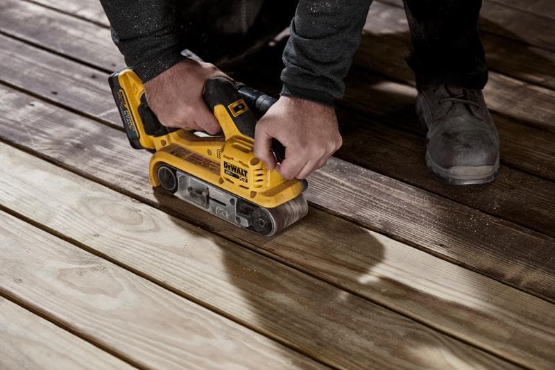 Uma pessoa utiliza uma lixadeira de cinta amarela sem fio DEWALT para lixar tábuas de madeira no chão.