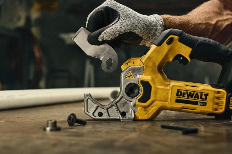 Uma pessoa usando uma luva de proteção segura um acessório de lâmina ao lado de uma ferramenta elétrica amarela DEWALT 20V, que está sobre uma bancada de trabalho. Pequenas peças de ferragem estão visíveis na mesa, com um tubo plástico branco ao fundo.
