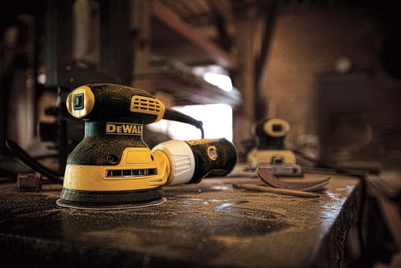 Close-up de uma lixadeira elétrica DEWALT DWE6421 sobre uma bancada empoeirada em uma oficina, com discos de lixamento e outra lixadeira visíveis ao fundo.