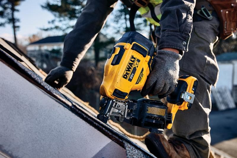 Uma pessoa usando luvas de trabalho opera uma pregadora sem fio amarela DEWALT para telhados em um telhado inclinado ao ar livre.