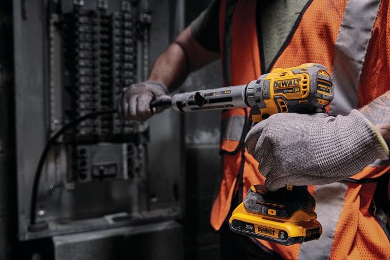Uma pessoa usando luvas e colete de segurança laranja utiliza uma ferramenta elétrica sem fio DEWALT 20V para trabalhar em um painel elétrico.