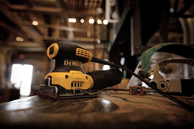 Uma lixadeira elétrica DEWALT DWE6411K está sobre uma bancada empoeirada em uma oficina, com o cabo conectado e equipamento de segurança visível ao fundo.