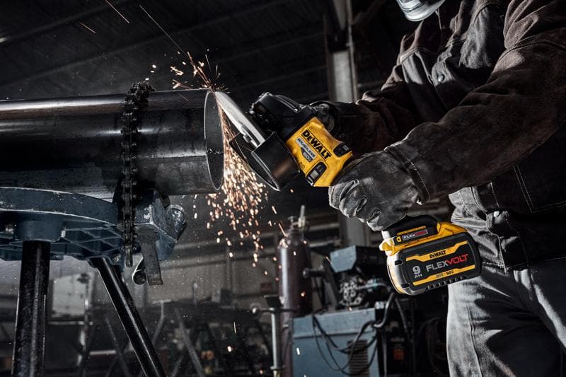 Uma pessoa usando equipamento de proteção utiliza uma esmerilhadeira angular sem fio DEWALT para cortar um grande tubo de metal em uma oficina industrial, com faíscas voando durante o corte.