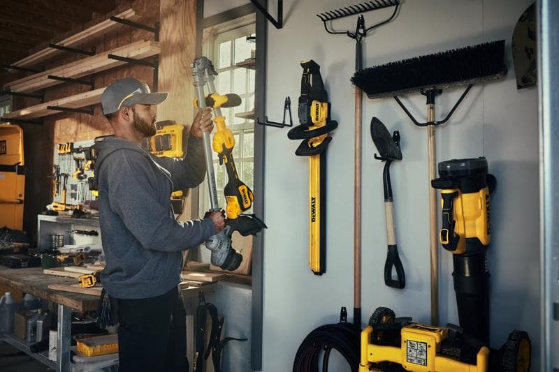 A person in a workshop holds a DEWALT DCST926 power tool. Various DEWALT tools and garden equipment are organized and hanging on the wall, with other tools and supplies visible on shelves and the workbench.