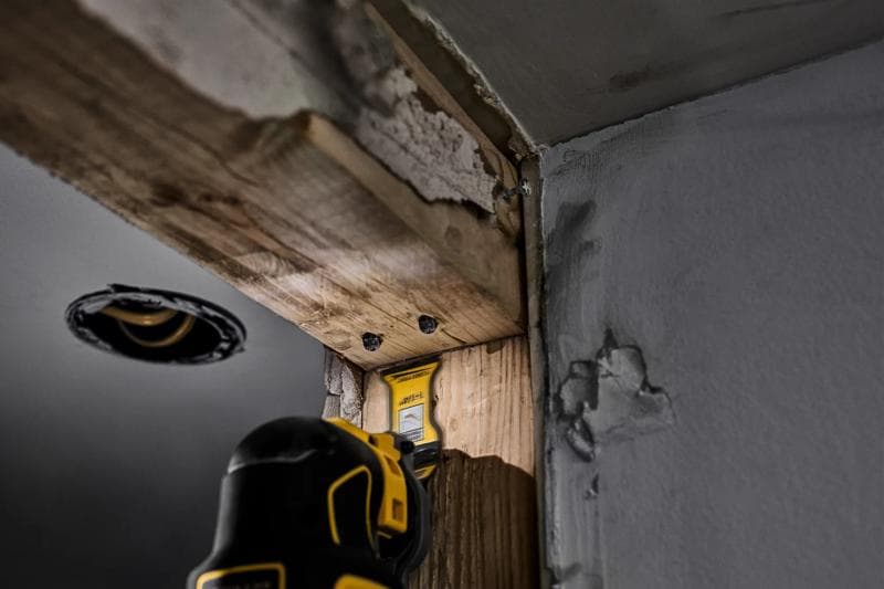 Uma ferramenta oscilante DEWALT sendo usada para cortar madeira em um canto de um local de construção, com drywall visível e madeira exposta.