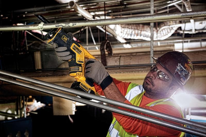 Uma pessoa usando luvas de proteção, colete de segurança e capacete está usando uma serra recíproca sem fio DEWALT DCS367B para cortar um tubo de metal em um ambiente industrial. O rosto da pessoa está desfocado para preservar a privacidade.