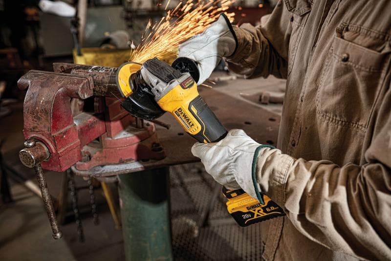Pessoa usando luvas de proteção opera uma esmerilhadeira angular sem fio DEWALT, modelo DCG413R2, para cortar ou lixar metal preso em uma morsa, com faíscas em uma oficina.