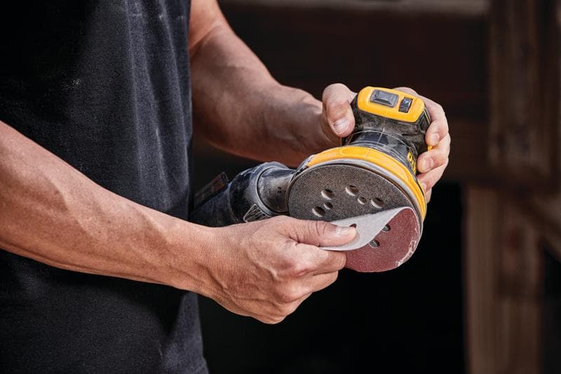 Uma pessoa segurando uma lixadeira orbital sem fio DEWALT, modelo DCW210B, fixando um disco de lixa na ferramenta.