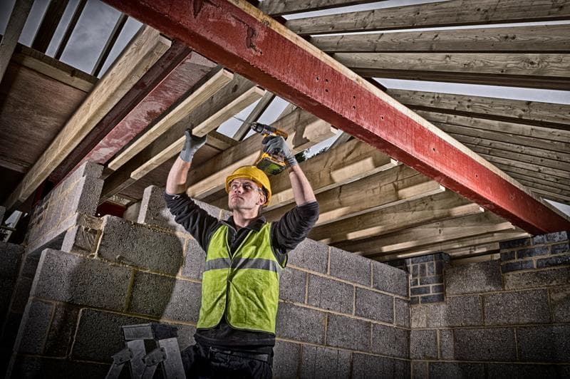 Um trabalhador da construção civil usando capacete amarelo e colete de alta visibilidade utiliza uma furadeira sem fio DEWALT para fixar vigas de madeira em uma viga de aço vermelha dentro de um edifício em construção. O rosto do trabalhador está desfocado.