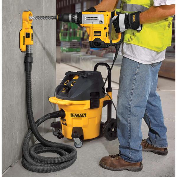 Um trabalhador da construção civil utiliza uma furadeira DEWALT e um sistema de extração de poeira para perfurar uma parede de concreto. O conjunto inclui um aspirador DEWALT conectado por uma mangueira à área de perfuração, reduzindo a poeira durante a operação.