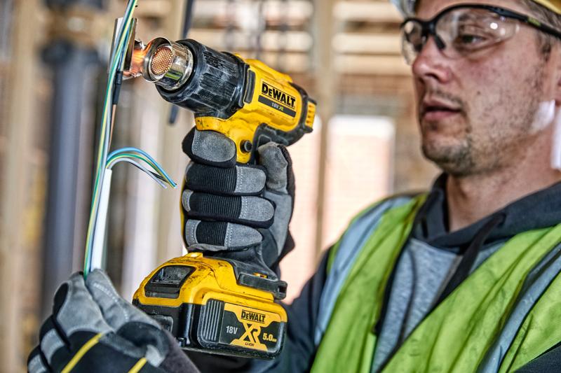 Uma pessoa usando luvas de trabalho e colete de segurança utiliza um soprador térmico sem fio DEWALT DCE530N para aplicar calor em fios elétricos em um canteiro de obras. O rosto da pessoa está borrado para preservar a privacidade.