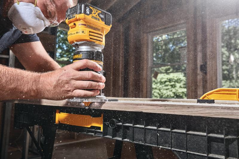 Close-up de uma pessoa usando uma tupia sem fio DEWALT para moldar a borda de uma tábua de madeira em um cavalete, com serragem voando e a pessoa usando máscara de proteção.
