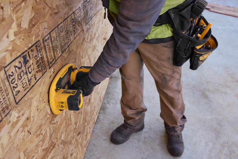 Um trabalhador usando calças marrons e luvas de proteção utiliza uma ferramenta portátil DEWALT em uma grande chapa de OSB em um canteiro de obras. O trabalhador possui um cinto de ferramentas com várias ferramentas manuais.