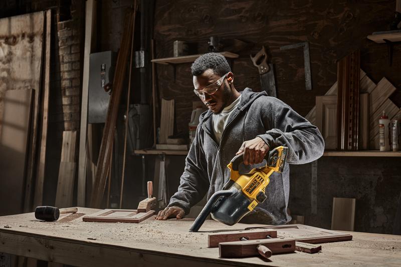 Uma pessoa está usando um aspirador portátil DEWALT modelo DCV501HB para limpar serragem de uma bancada em uma oficina de marcenaria. Diversas ferramentas e peças de madeira são visíveis na mesa e nas prateleiras ao fundo.