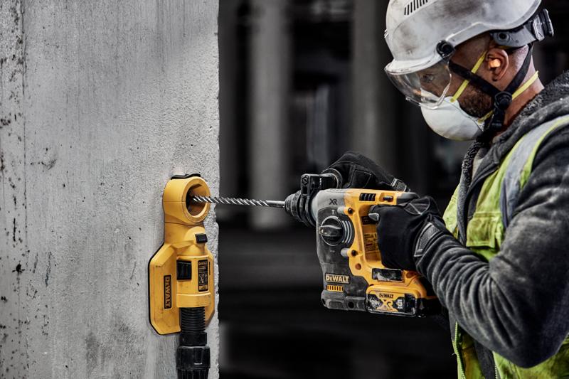 Um trabalhador da construção usando equipamento de segurança está utilizando uma furadeira elétrica amarela da DEWALT para perfurar uma parede de concreto. Um acessório de extração de poeira DEWALT está conectado à parede para coletar detritos.