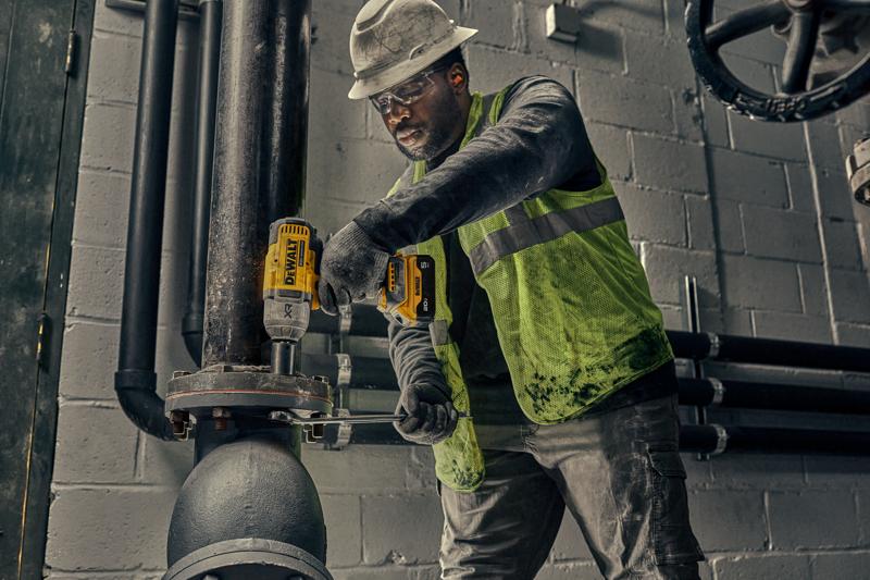 Um trabalhador com capacete e colete de segurança usa uma ferramenta elétrica amarela DEWALT para apertar parafusos em um grande tubo industrial dentro de uma instalação.