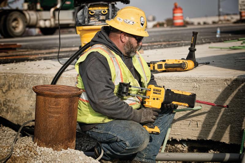 Um operário da construção usando capacete amarelo, colete de segurança e luvas está utilizando uma ferramenta elétrica DEWALT para perfurar concreto em um canteiro de obras ao ar livre. O rosto do trabalhador está borrado para preservar a privacidade. Outras ferramentas DEWALT e equipamentos de construção aparecem ao fundo.