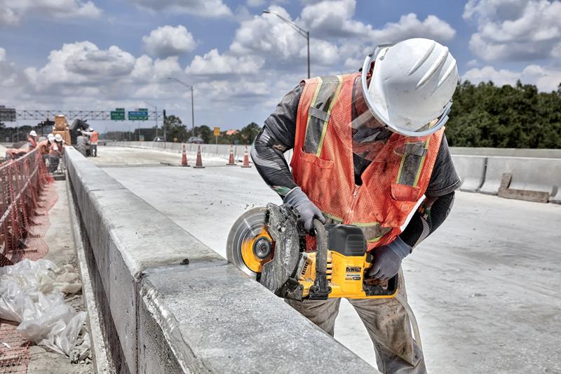 Um trabalhador da construção civil usando capacete branco e colete de segurança laranja está utilizando uma serra sem fio DEWALT (modelo DCS692B) para cortar uma barreira de concreto em um canteiro de obras.