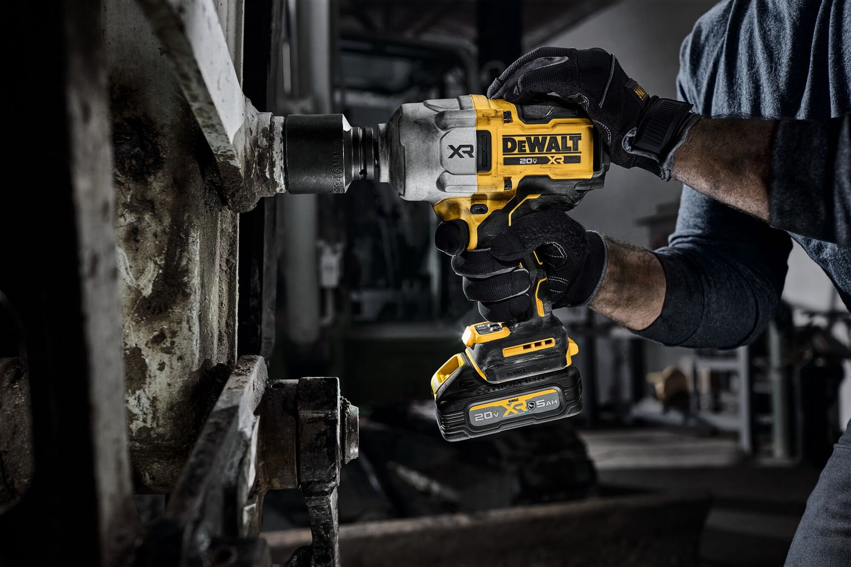Mãos com luvas usam uma chave de impacto DEWALT sem fio em parafuso metálico em ambiente industrial.